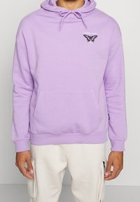 Lavendelfärgad hoodie med en framficka, dragsko i huvan och svart grafisk detaljering. Tillverkad av mjukt tyg, med ribbade ärmslut och nederkant.