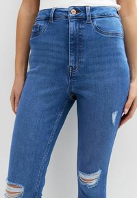 Vaqueros de mezclilla azul de cintura alta con un diseño desgastado. Presenta detalles rasgados en las rodillas y un botón de cobre en la cintura.