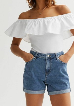 Femme portant un haut blanc à volant et épaules dénudées ainsi qu’un short en jean bleu, les mains dans les poches, sur fond uni.