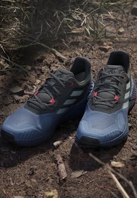 Niebieskie i czarne buty sportowe z siateczkowymi cholewkami, gumową podeszwą i czerwonymi akcentami. Obejmuje trzy odblaskowe paski i okrągły przód.