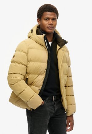 Chaqueta tipo pluma en beige claro, con capucha desmontable, acolchado cosido y acentos negros en la cremallera y el forro interior.