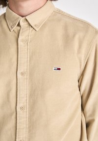 Camicia in corduroy beige con colletto a punta e bottoni frontali. Presenta un piccolo logo ricamato in blu navy, bianco e rosso. Morbida al tatto.