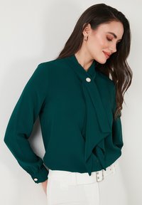 Teal blouse featuring a tied collar, long sleeves, and a button closure, made from smooth fabric with subtle sheen. Accented with a decorative button.