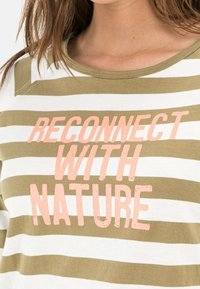 Femme portant un t-shirt rayé blanc et vert olive avec un texte rose disant "RECONNECTEZ AVEC LA NATURE" sur la poitrine.
