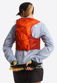 Pomarańczowy plecak do biegania z siateczkowymi akcentami i regulowanymi paskami, w zestawie z lekką szarą kurtką i czarnymi szortami. Trzyma kijki trekkingowe.