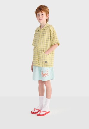 Jong roodharig kind met een geel geruit kort overhemd, lichtblauwe korte broek met oranje print, witte sokken en rode slippers.