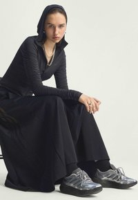 Black zip-up top with textured sleeves, paired with a flowing black skirt. Silver athletic shoes with mesh detailing and black soles.