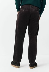Person wearing dark brown corduroy pants and black shoes, standing with hands in pockets against plain light background.