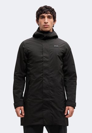 Jeune homme portant une longue veste à capuche noire avec le logo Helly Hansen sur la poitrine, debout devant un fond clair uni.