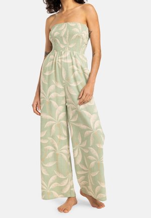 Kvinde iført en skulderfri jumpsuit med brede ben med et grønt og beige palmebladsmønster, stående barfodet mod en hvid baggrund.
