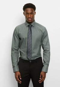 Dunkelgrünes, langärmeliges Hemd mit spitzem Kragen, kombiniert mit einer dunklen, gestreiften Krawatte und schwarzen Hosen. Glatter Stoff und taillierte Passform.