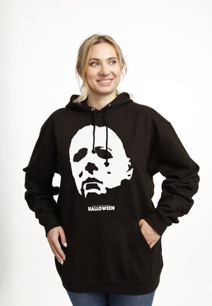 Uśmiechnięta kobieta w czarnej bluzie z kapturem, nosząca dużą białą maskę Michaela Myersa z napisem "John Carpenter's Halloween", z rękami w kieszeniach.