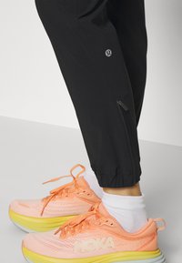 Pantaloni sport negri cu mansete la gleznă cu fermoar, asortați cu pantofi de alergare portocalii și galbeni, având un superior din plasă și o talpă groasă.