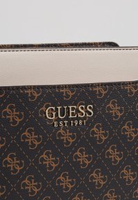 Černá texturovaná kožená peněženka s hnědým vzorem loga, se smetanovým akcentním panelem a zlatým logem značky "GUESS EST 1981."