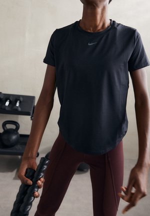 Personne portant un t-shirt Nike noir et des leggings bordeaux tenant une barre de fitness noire texturée dans un espace d'entraînement avec des poids en arrière-plan.