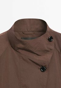 Brun jackkrage med en unik asymmetrisk design, som har en svart knapp och textil med subtila ränder. Minimalistisk stil.