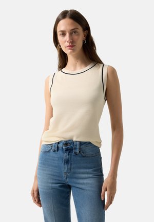 Femme portant un haut en tricot crème sans manches avec un bord noir et un jean bleu taille haute, posant devant un fond blanc.