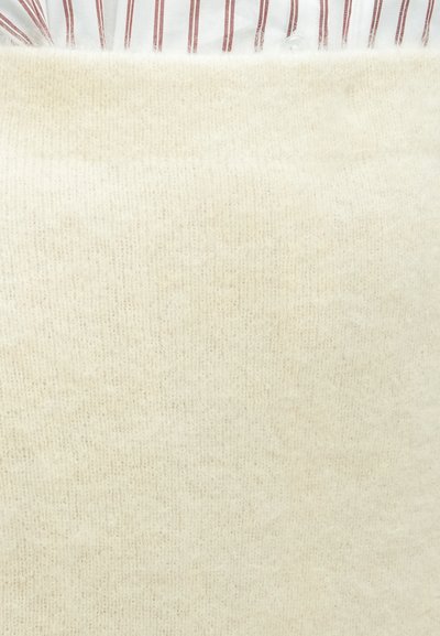 Asmuo dėvi kreminės spalvos pūkuotą megztinį ant baltos su raudonomis vertikaliomis juostelėmis marškinių, parodytas viršutinės liemens dalies artimas vaizdas.