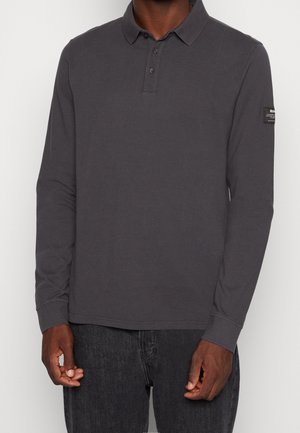 Homme portant un polo gris foncé à manches longues avec col boutonné et un jean noir, se tenant debout devant un fond clair uni.