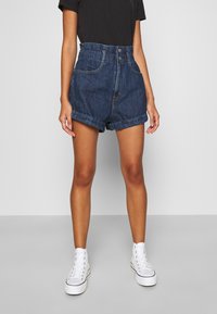 Persona vestida con pantalones cortos de mezclilla de tiro alto y dobladillo enrollado, camiseta negra y zapatillas deportivas blancas de caña alta, de pie contra un fondo plano.