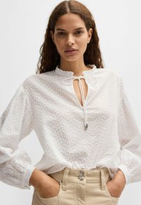 Blusa bianca con texture, colletto arricciato e chiusura con laccio davanti. Maniche lunghe con polsini elastici. Abbinata a pantaloni beige.