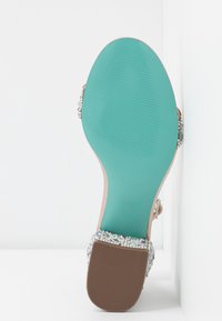Talons d'une sandale avec une semelle turquoise, des brides beiges et une bande décorative de strass clairs sur le bout. Surface texturée.