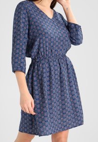 Robe bleue à motifs avec un décolleté en V, des manches trois quarts et une taille élastique. Le tissu présente un motif géométrique en rouge et blanc.