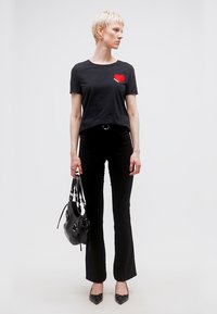 Νεαρό άτομο με κοντά ξανθά μαλλιά που φοράει μαύρο T-shirt με κόκκινη καρδιά, μαύρο παντελόνι, μαύρες γόβες και κρατάει μια μαύρη τσάντα χειρός.