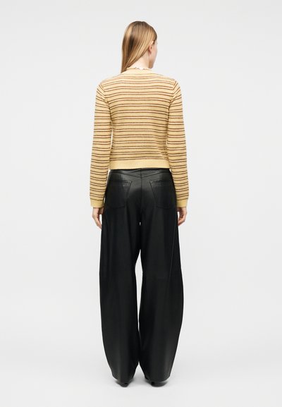 Γυναίκα με μακριά ξανθά μαλλιά, ντυμένη με πουλόβερ με κίτρινες και καφέ ρίγες και φαρδιά μαύρα δερμάτινα παντελόνια, στέκεται γυρισμένη προς τα πίσω.