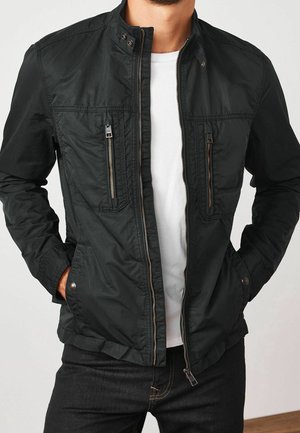 Homme portant une veste noire dézippée avec des poches sur la poitrine et les côtés, par-dessus un t-shirt blanc et un jean foncé, les mains dans les poches inférieures.