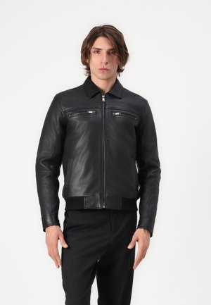 Junger Mann steht frontal, trägt eine schwarze Lederjacke mit Reißverschluss und Reißverschlusstaschen auf der Brust sowie schwarze Hosen vor weißem Hintergrund.