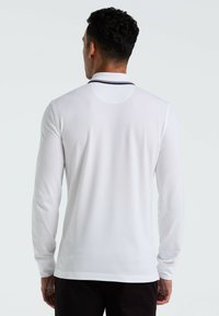 Man met kort krullend haar die een witte langermige poloshirt met zwarte biezen op de kraag draagt, met zijn rug naar de camera gekeerd tegen een effen achtergrond.
