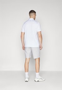 Hvit polo-shirt med korte ermer, kombinert med lysegrå shorts. Har en subtil logo på øvre del av ryggen og sporty fottøy.