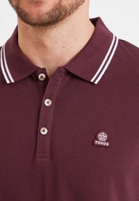 Burgundy polo shirt with a textured fabric, white striped collar, three button placket, and a circular TOG24 logo on the chest.