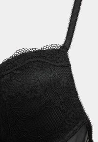 Zwarte kant bralette met bloemenkant overlay, gestructureerde cups en verstelbare bandjes met geschulpte randen voor extra detail.