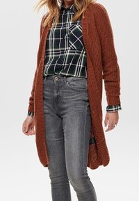 Vrouw in een roestkleurige gebreide cardigan over een groen en marineblauw geruite blouse, ingestopt in grijze highwaisted skinny jeans.