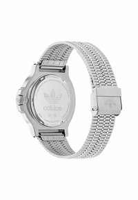 Sudraba nerūsējoša tērauda pulkstenis ar sietveida jostu, apaļa korpusa un teksturēta aizmugure ar izreljefētu logo. Iekļauta sprādze.