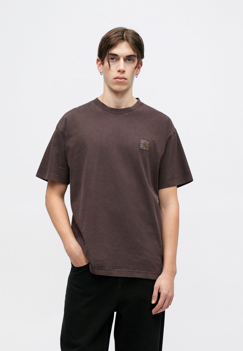 Braunes Baumwoll-T-Shirt mit kurzen Ärmeln, Rundhalsausschnitt und einem kleinen Logopatch auf der linken Brust. Das Modell steht mit Händen in den Taschen.
