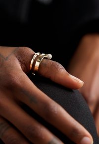 Gold-colored metal rings stack on a finger, featuring a smooth band, a textured ball accent, and a narrow band with small clear stones.