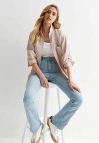 Gestreepte button-up blouse, lichtroze en wit. Hoogwaardige lichtblauwe jeans. Witte sneakers met tan accenten. Zittend op een witte kruk.