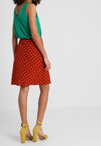 Zielony top bez rękawów w połączeniu z czerwoną spódnicą w kropki. Żółte sandały na obcasie z paskami na kostki. Gładki materiał z płynącą sylwetką.