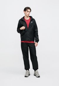 Fiatal férfi fekete Nike dzsekiben és nadrágban, piros pólóban, szürke tornacipőben, fekete keresztpántos táskát viselve, fehér háttér előtt áll.