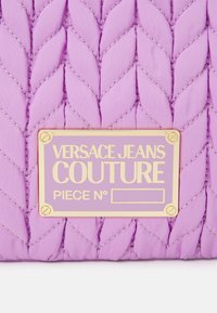 Borsa a tracolla in tessuto trapuntato viola con un motivo intrecciato e accessori in oro. Presenta una placca dorata con l'iscrizione "VERSACE JEANS COUTURE".