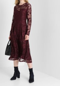Bordó krajková šaty s dlouhými rukávy, květinovým vzorem, plisovanou sukni, nošené s černými kotníkovými botami a držící černou kabelku.