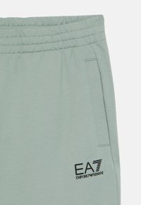 Gai zaļi sporta bikses no mīksta auduma, ar elastīgu jostu, sānu kabatu un melnu EA7 Emporio Armani logo priekšpusē.