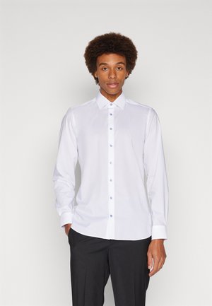 Mann mit Afro-Frisur, der ein weißes Hemd mit langen Ärmeln und schwarze Hose trägt, steht vor einem schlichten hellen Hintergrund.