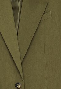 Blazer verde oliva con revers nota, chiusura a un bottone e taschino sul petto. Il tessuto ha una consistenza liscia con una leggera lucentezza.