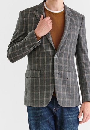 Man draagt een grijze geruite blazer over een bruine trui en blauwe jeans, stelt de blazer kraag bij met zijn linkerhand.