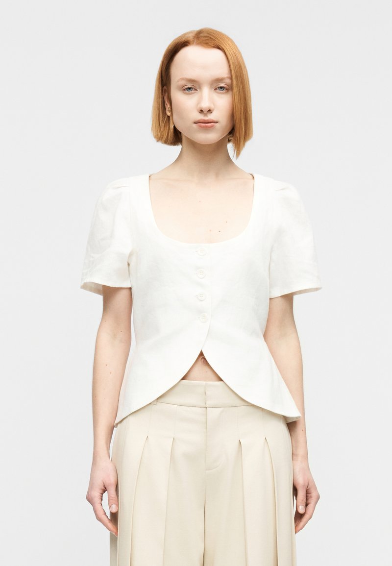 Λευκό λινο πουκάμισο με κοντά φουσκωτά μανίκια, τετράγωνη λαιμόκοψη και κούμπωμα εμπρός με πέντε κουμπιά. Κοντό σχέδιο με στρογγυλεμένο τελείωμα.