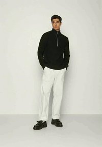 Melns džemperis ar rāvējslēdzēju, izgatavots no strukturētas adīšanas, kombinēts ar baltām, brīvām biksēm. Modelis nēsā melnas kurpes ar gludu virsmu.